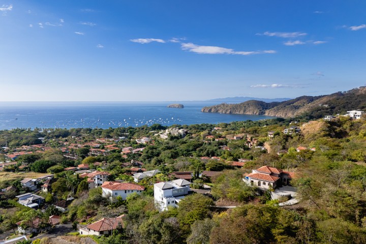 Aerial view of a coastal town with red-roofed houses, dense greenery, and blue ocean under a clear sky.