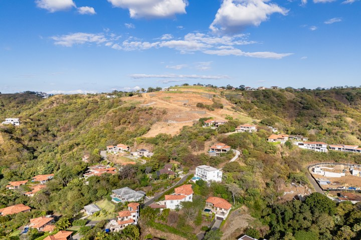 Aerial view of hilly terrain with houses scattered among trees under a partly cloudy sky.