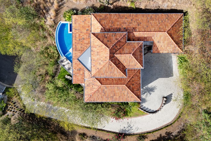 Aerial view of a house with a red-tiled roof, curved driveway, and a blue pool surrounded by greenery.