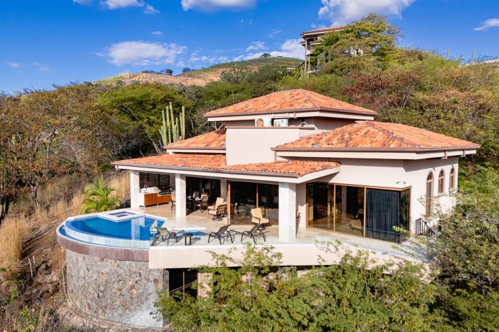 Hilltop villa with terracotta roof, infinity pool, and surrounding trees under a blue sky.