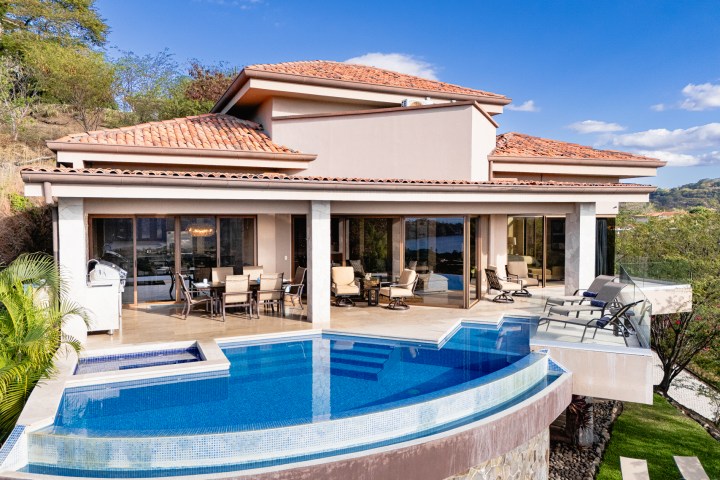 Luxury villa with terracotta roof, glass doors, and elevated infinity pool overlooking a scenic landscape.