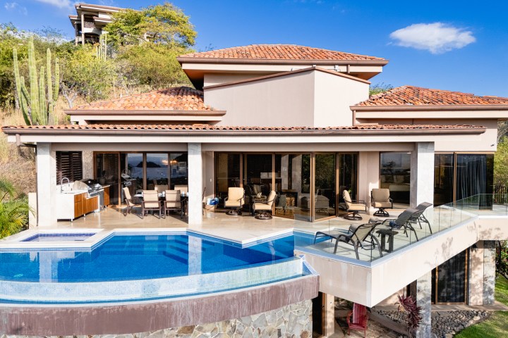 Modern house with pool, patio furniture, and glass railings on a sunny day.