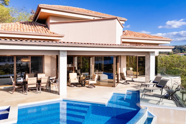 Luxurious villa with patio, outdoor seating, and infinity pool under a clear blue sky.