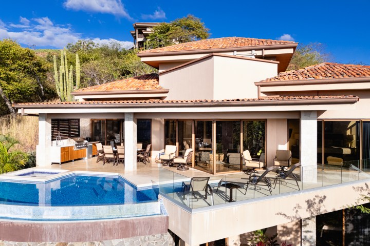 Modern villa with glass walls, infinity pool, outdoor seating, and terracotta roof under a clear blue sky.