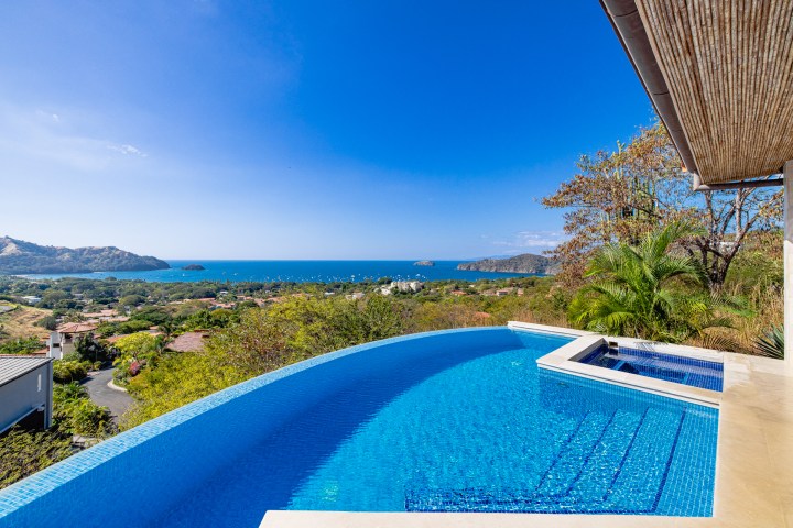 Infinity pool with ocean view, surrounded by greenery and hills under a clear blue sky.