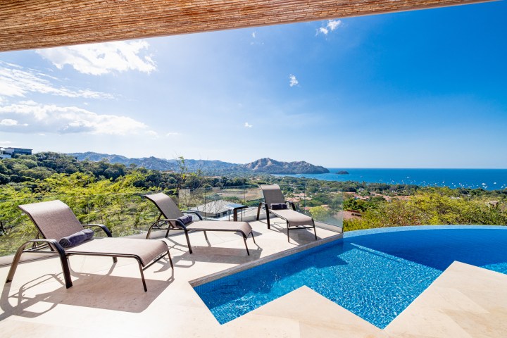 Outdoor loungers by an infinity pool overlooking a scenic ocean view with mountains.