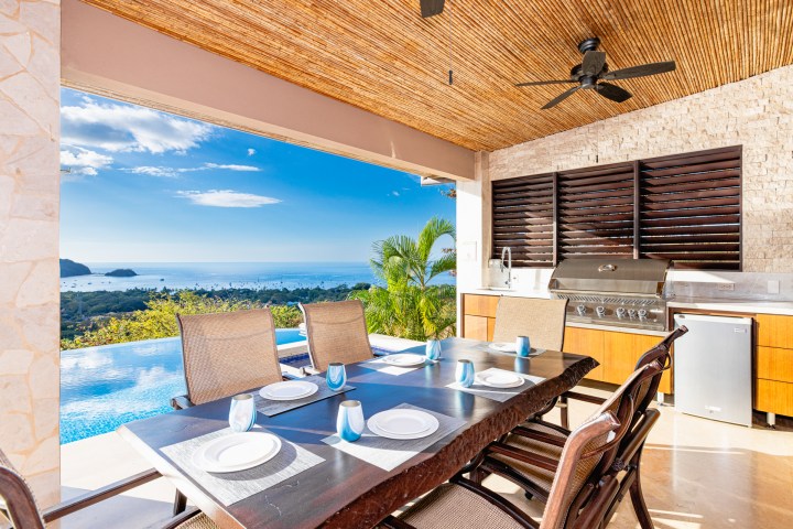 Outdoor dining area with wooden table, sea view, and grill under a bamboo ceiling.
