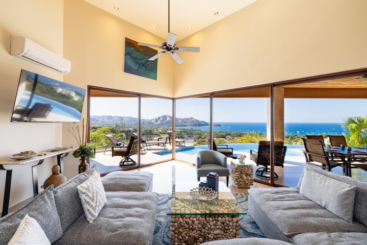 Spacious living area with ocean view, gray sofas, glass table, TV, and outdoor seating by pool.