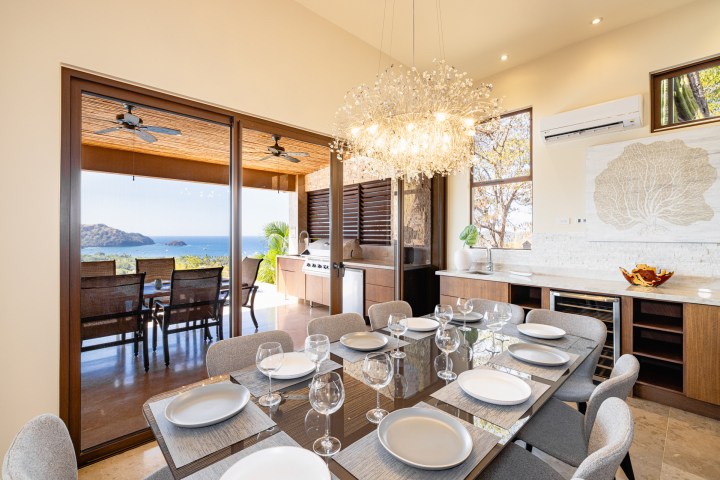 Elegant dining area with set table and chandelier, leading to outdoor patio with ocean view.