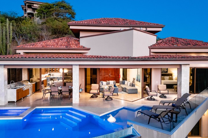 Luxurious villa with red-tiled roof, outdoor pool, and patio under a sunset sky.