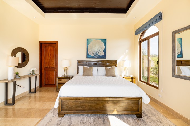 Elegant bedroom with wooden bed, white bedding, large windows, and wall art of sea coral.