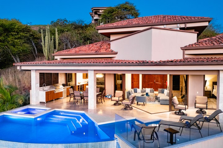 Luxury villa with open patio, pool, and outdoor furniture at dusk, surrounded by greenery.