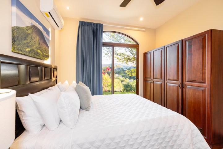 Bedroom with a large bed, wooden wardrobe, and window view of greenery.