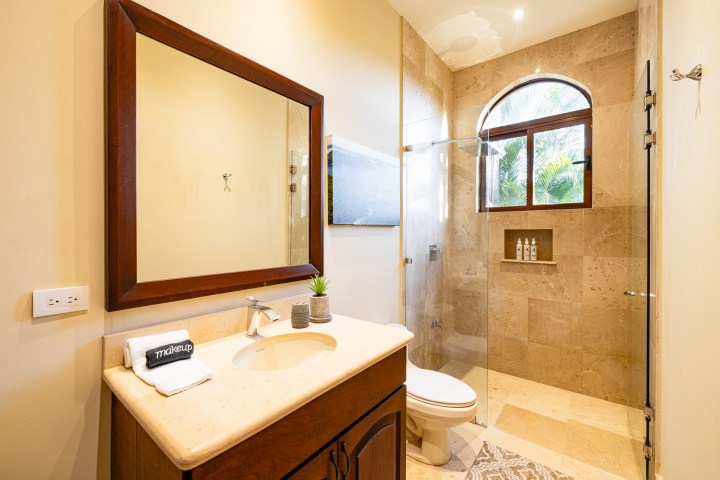 Bathroom with a wooden vanity, mirror, glass shower, and arched window with plants outside.