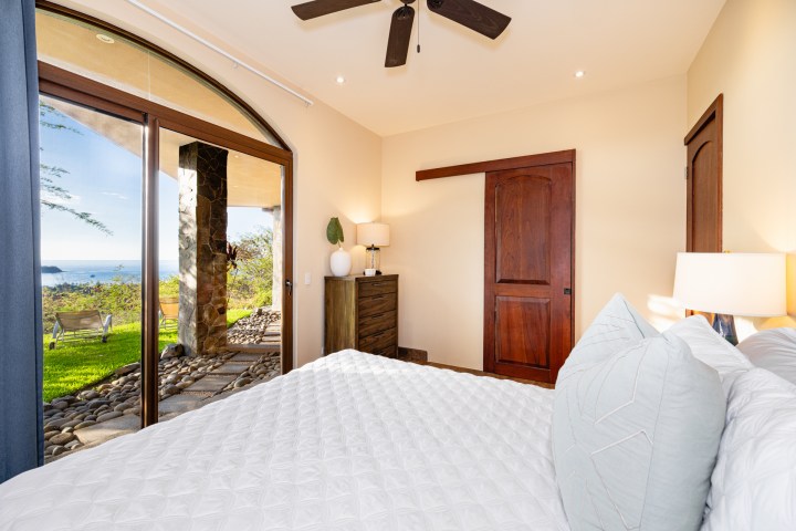 Bedroom with white bedding, brown furniture, glass doors open to grassy patio and ocean view.