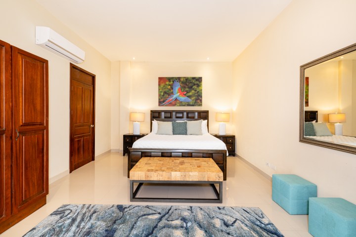 Bedroom with a bed, wooden doors, a painting, lamps, bench, mirror, and two blue ottomans.