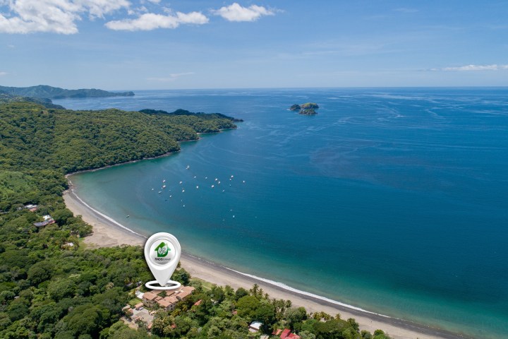 Aerial view of a beach with green coastline, blue ocean, and a location pin marker on a building.