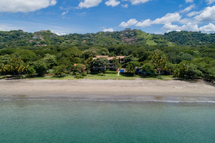 Beachfront view with trees, buildings, hills, and blue sky with clouds.