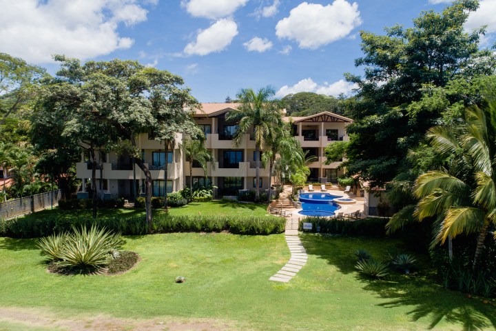 Apartment building with pool, surrounded by trees and lush greenery under a partly cloudy sky.