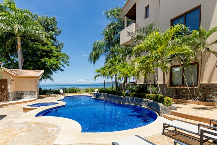 A tropical villa with a blue pool, palm trees, and ocean view under a clear sky.