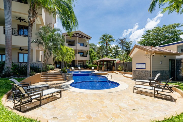 Outdoor pool area with lounge chairs, palm trees, and adjacent buildings under a clear blue sky.