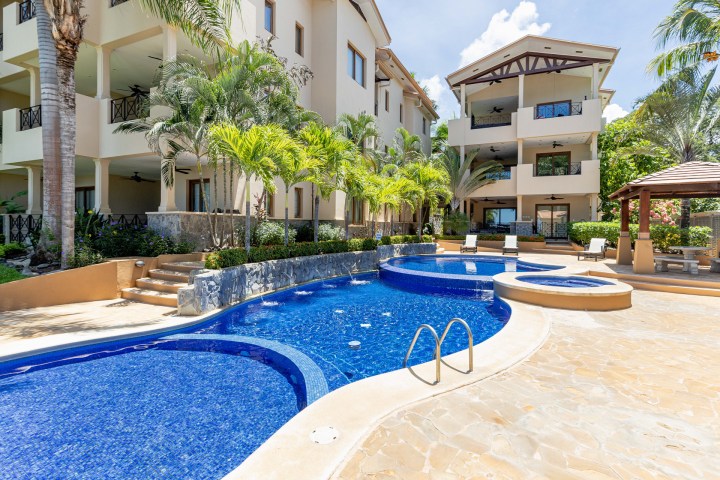 Outdoor swimming pool with curved design beside a multi-story beige building and palm trees.