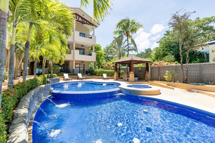Outdoor pool area with spa, tropical plants, and a modern building in the background.