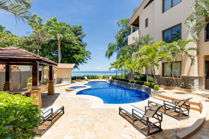 Tropical poolside area with loungers, palm trees, and ocean view under a clear blue sky.