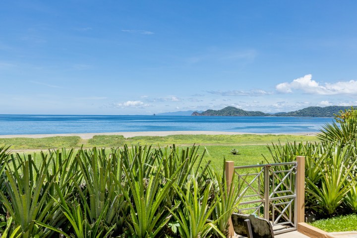 A gate overlooks a beach with green plants, ocean, and mountains under a clear blue sky.