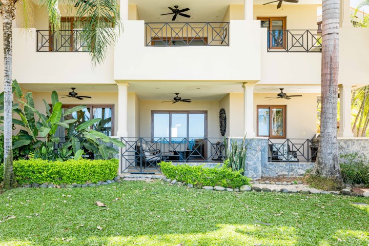 Two-story building with balconies, green plants, and ceiling fans, viewed from a grass lawn.