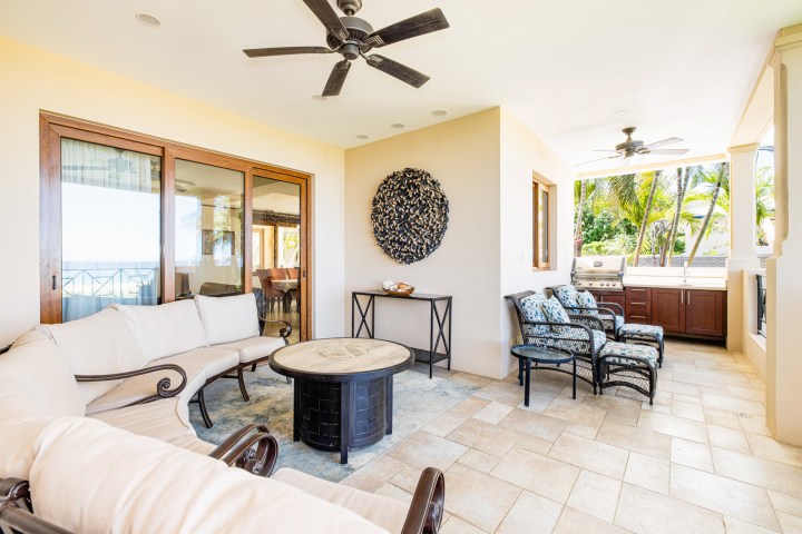 Patio with seating, round table, ceiling fans, and grill, overlooking palm trees.