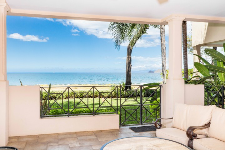 Beachfront patio with wicker furniture, ocean view, palm trees, and blue sky.