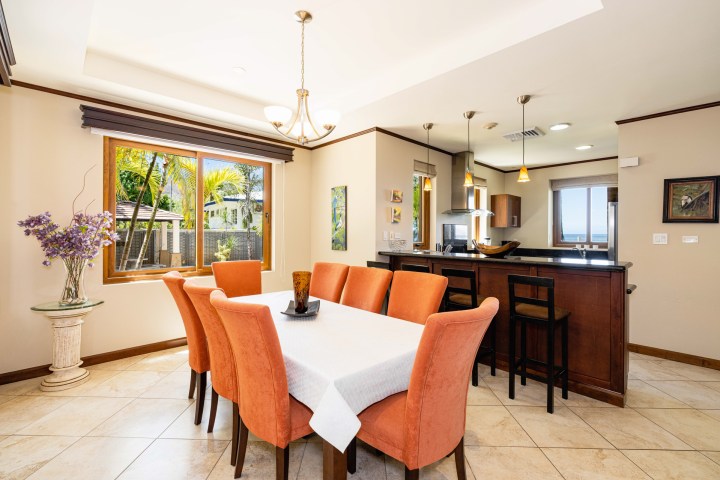 Modern dining room with orange chairs, white tablecloth, chandelier, and large window overlooking greenery.