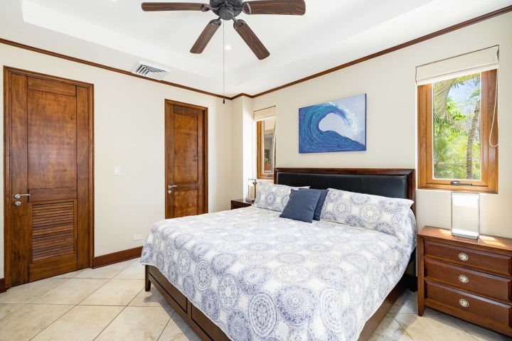 Bedroom with king bed, ceiling fan, ocean artwork, two wood doors, and windows with garden view.