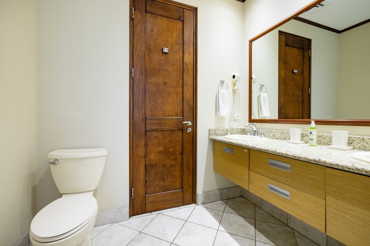 Modern bathroom with wooden door, toilet, double sink, large mirror, and tiled floor.