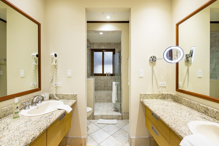 Bright bathroom with granite counters, two sinks, a mirror, and a door leading to a shower area.