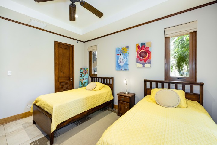 Bedroom with two yellow-covered twin beds, artwork, a fan, and a window overlooking greenery.