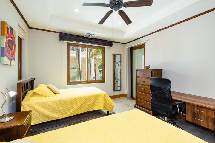 Bedroom with two yellow beds, ceiling fan, window, desk with chair, and dresser.