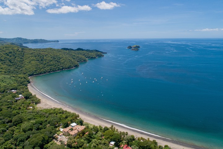 Aerial view of a coastal bay with boats, lush greenery, and distant hills under a blue sky.