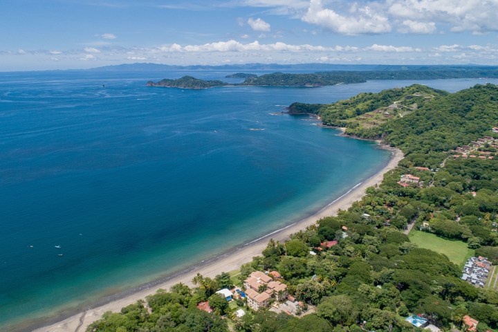Aerial view of a coastline with blue ocean, sandy beach, and lush green hills on a sunny day.