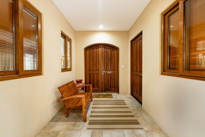 Hallway with wooden doors, two windows, a small bench, a ceramic pot, and a striped rug on tiled floor.