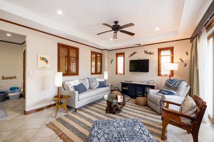 Bright living room with sofas, a ceiling fan, and a wall-mounted TV. Coastal decor and large windows add charm.