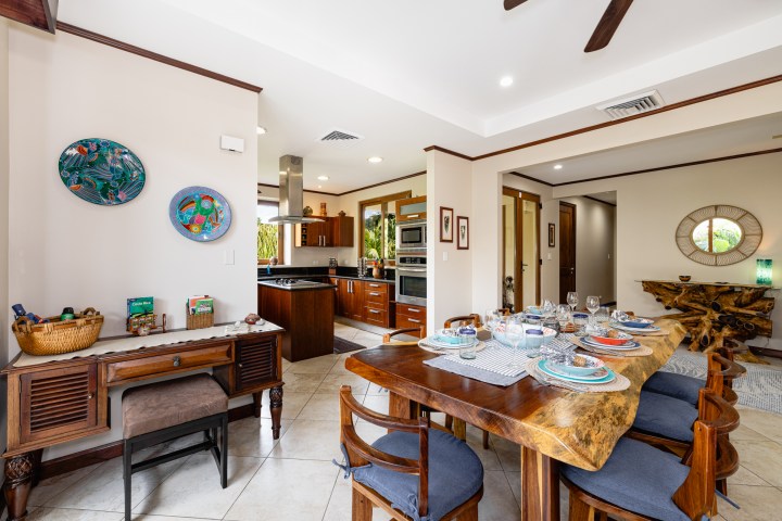 Modern dining room with wooden table, chairs, wall art, and view of kitchen with double ovens.