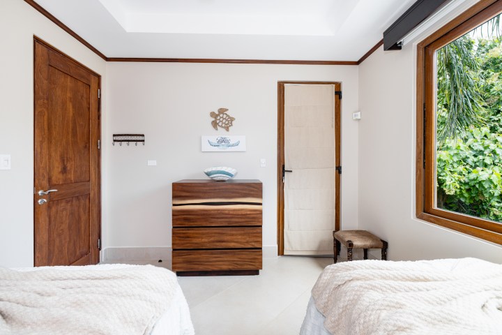 Bedroom with wooden dresser, artwork on wall, window view of greenery, and two beds with white covers.