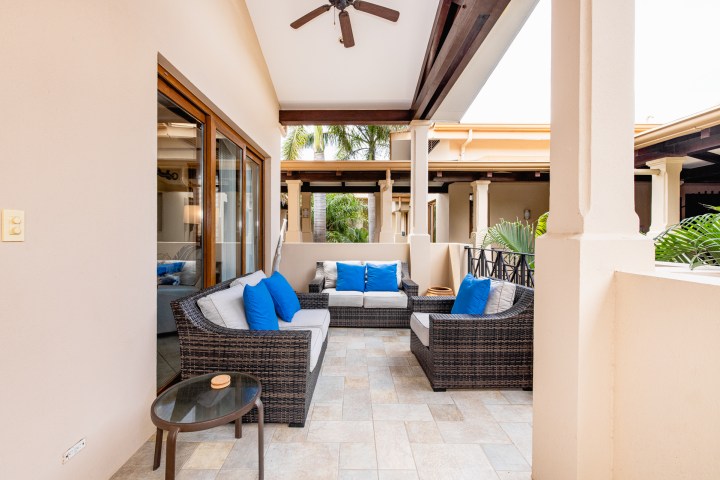 Outdoor patio with wicker sofas, blue cushions, a glass table, and ceiling fan under a covered area.