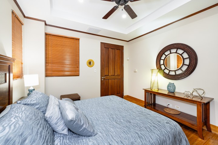 Bedroom with blue bedding, wooden furniture, round mirror, and ceiling fan.