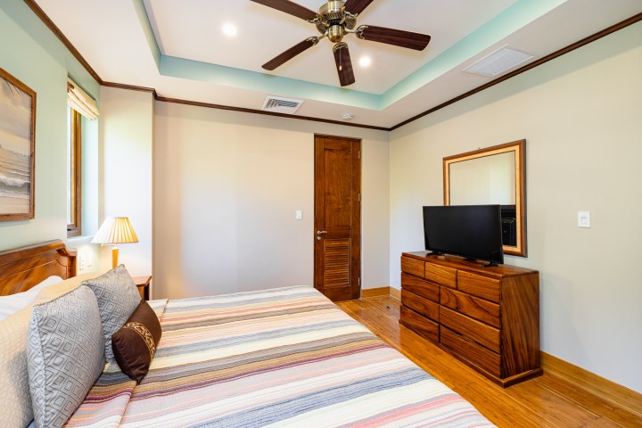 Bedroom with bed, striped bedding, wooden dresser, TV, ceiling fan, and lamp.