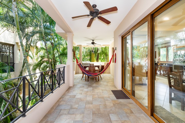 Outdoor patio with hammock, ceiling fans, dining table, surrounded by tropical plants and glass doors.