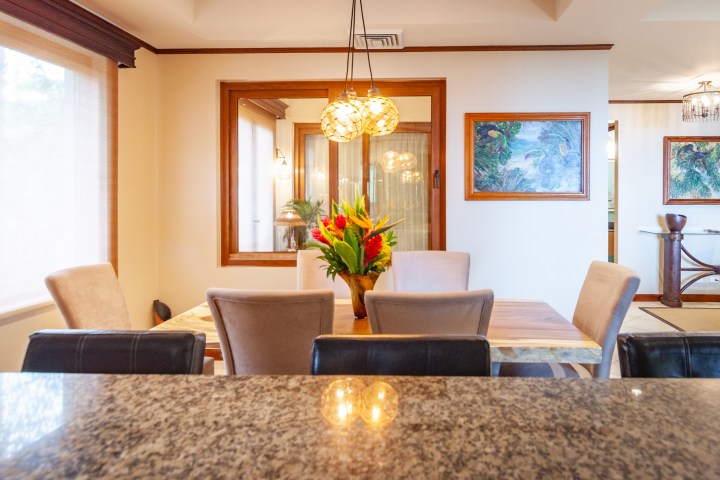 Dining area with table, chairs, pendant lights, and wall art, viewed from a granite counter.