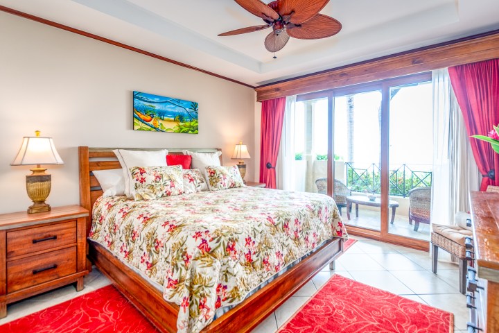 Bright bedroom with floral bedding, two lamps, ceiling fan, and balcony view with chairs.